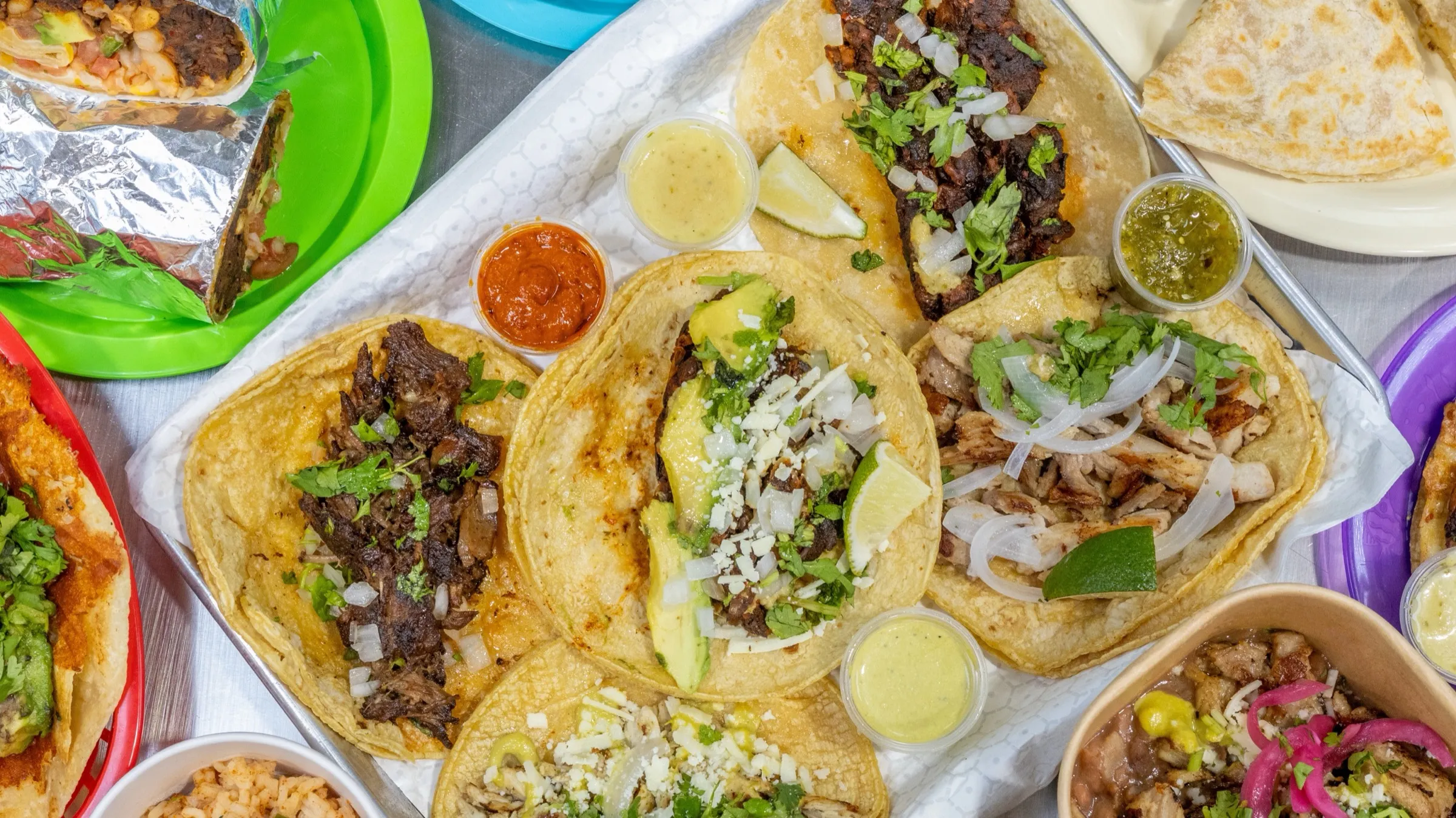 Overhead spread of tacos, burritos, bowls, sides and salsa cups on colorful plates