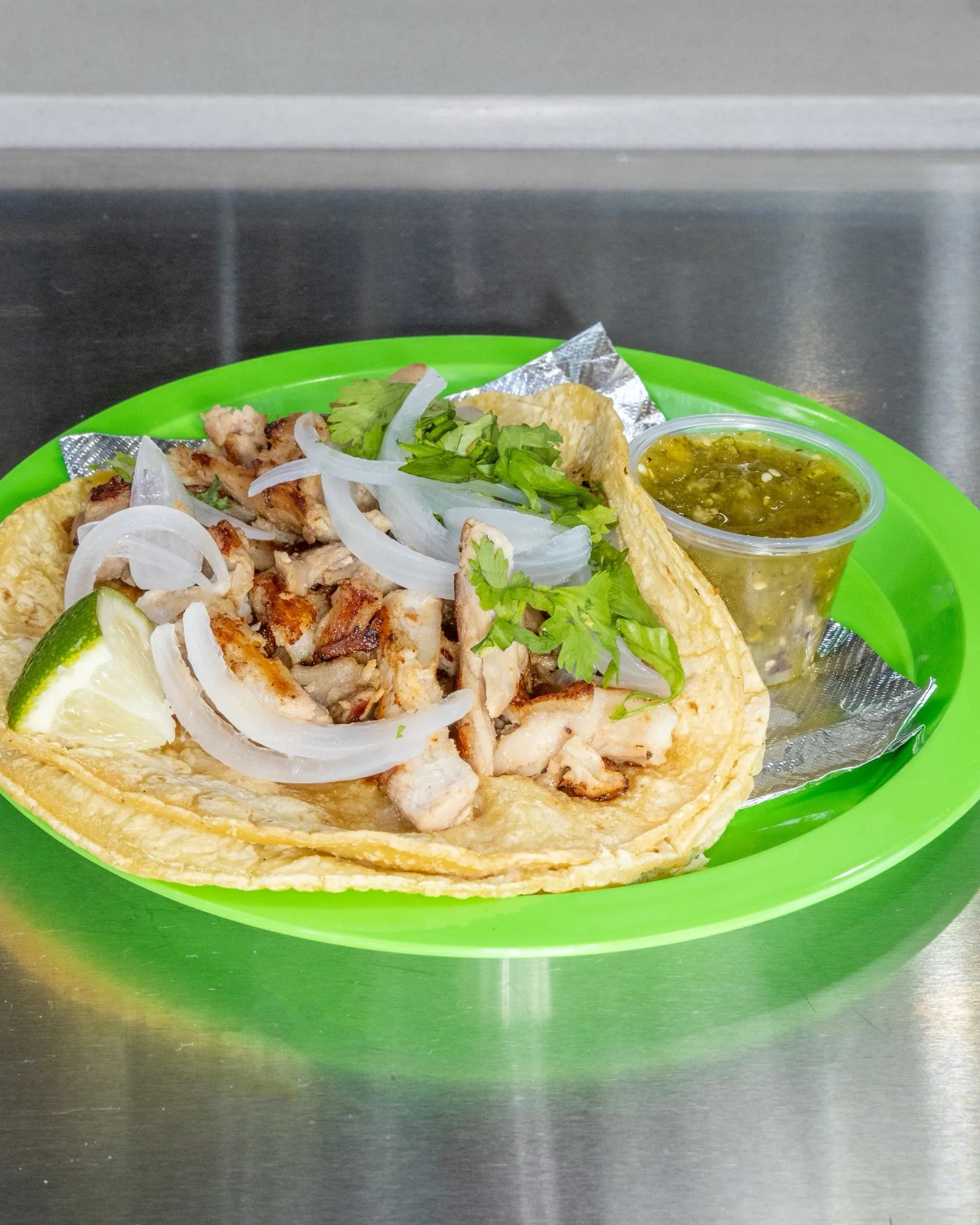 Two corn tortilla tacos topped with onion and cilantro on a green plate, lime and green salsa
