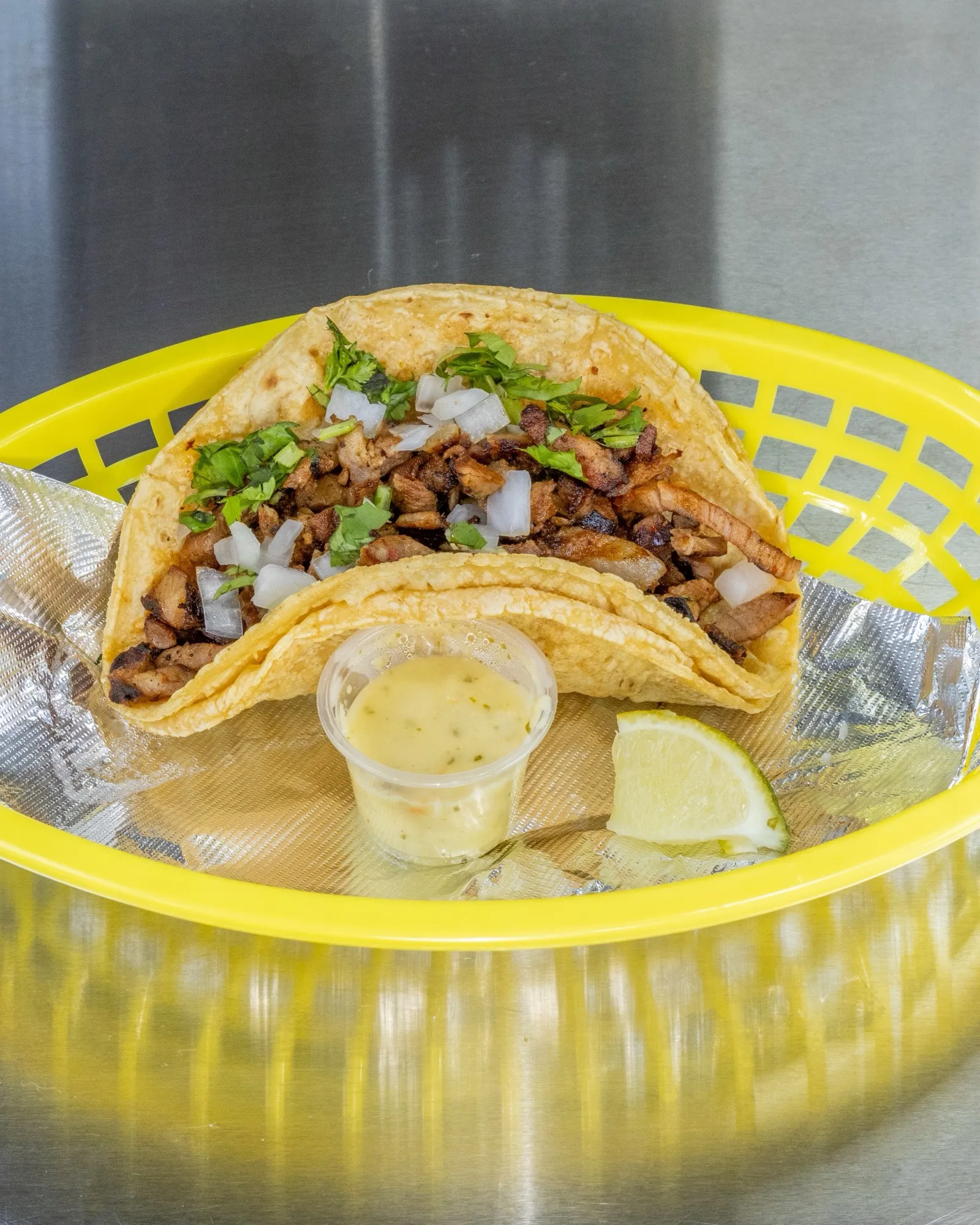 Two barbacoa tacos with onion and cilantro in a yellow basket, lime and salsa cup